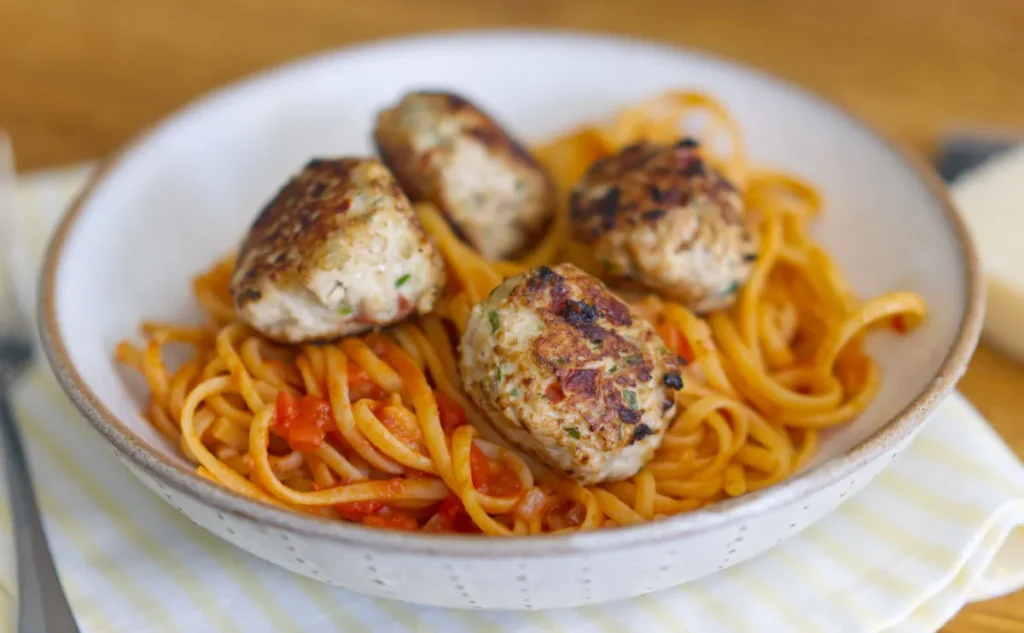Turkey meatballs on top of tomato and red pepper spaghetti, in a white bowl on top of a yellow and white striped tea towel.