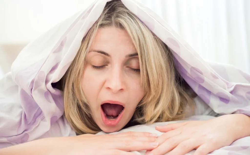 Tired woman yawning under a duvet, just her head is peeping out.