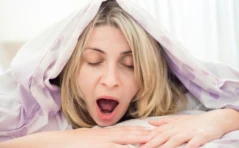 Tired woman yawning under a duvet, just her head is peeping out.