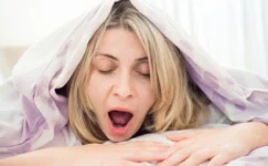 Tired woman yawning under a duvet, just her head is peeping out.