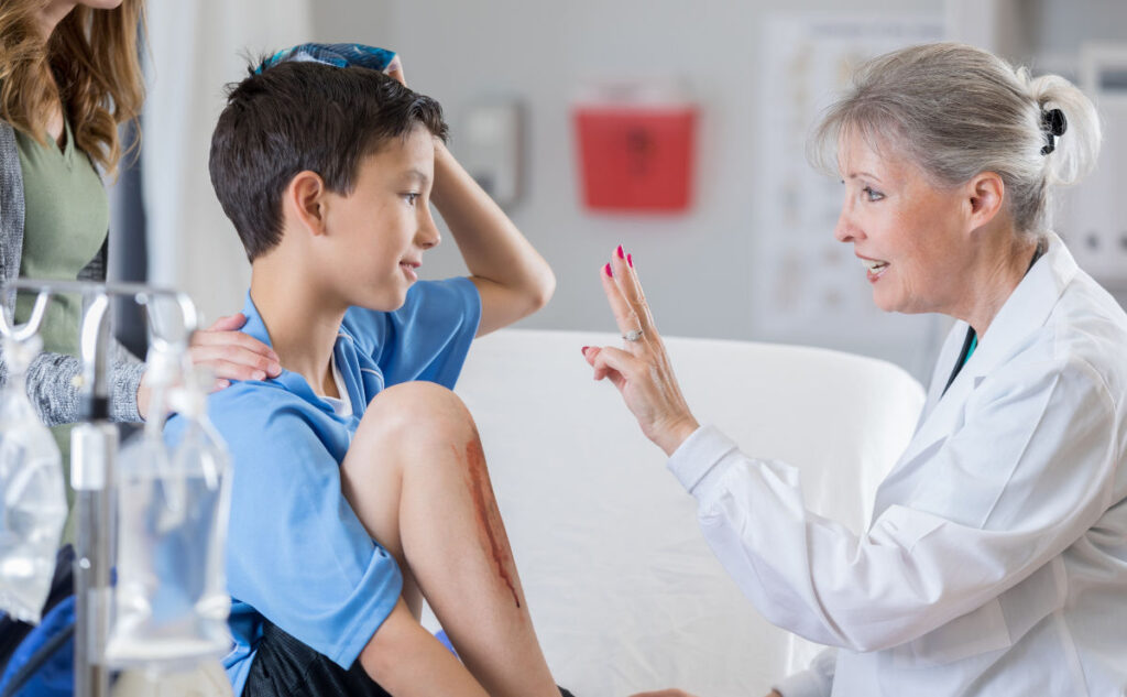 Female senior A&E doctor holds up two fingers to check the vision of an injured football player. The patient is holding an ice pack on his head. His mother is comforting him.