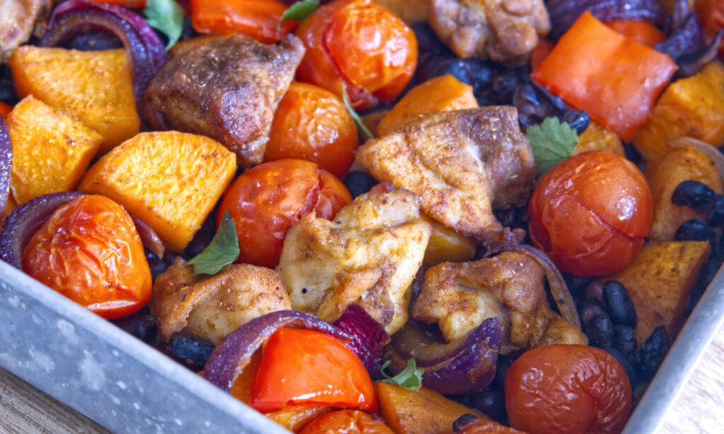 A close-up of a baking tray filled with roasted chicken, cherry tomatoes, red onions, sweet potatoes, and bell peppers, all cooked to a golden brown and garnished with fresh herbs.