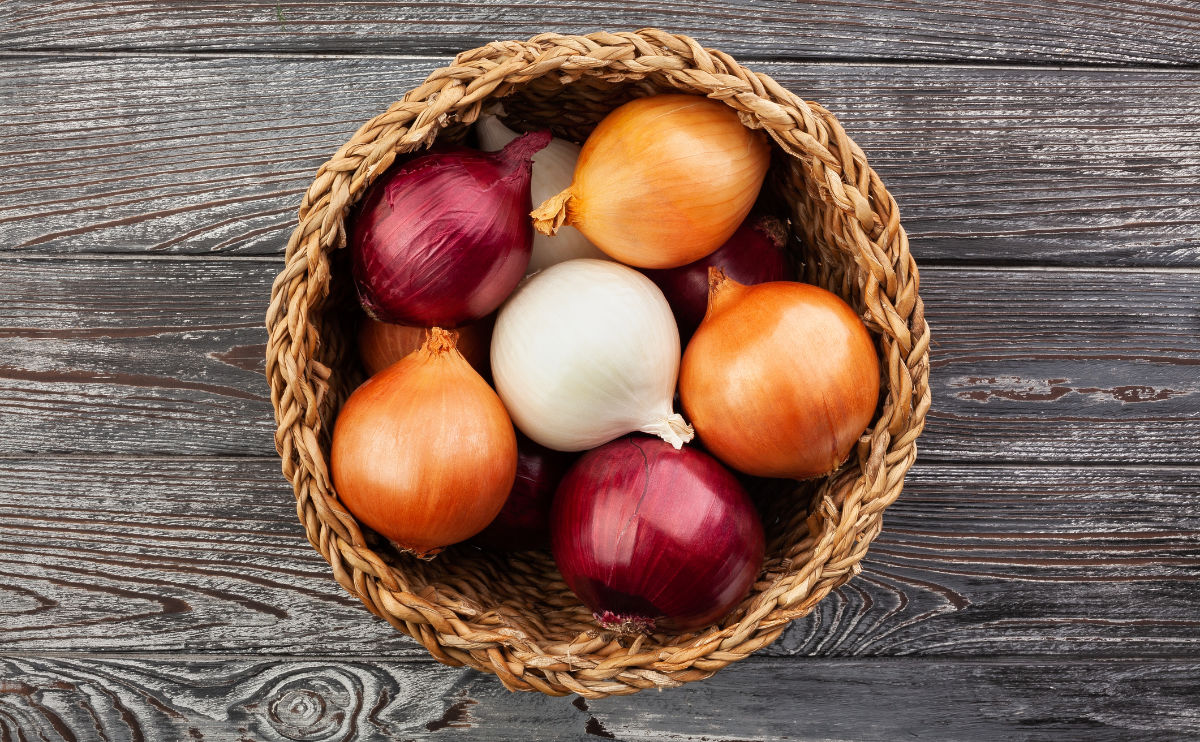 A woven basket filled with whole brown, yellow, red, and white onions sits on a rustic wooden surface.