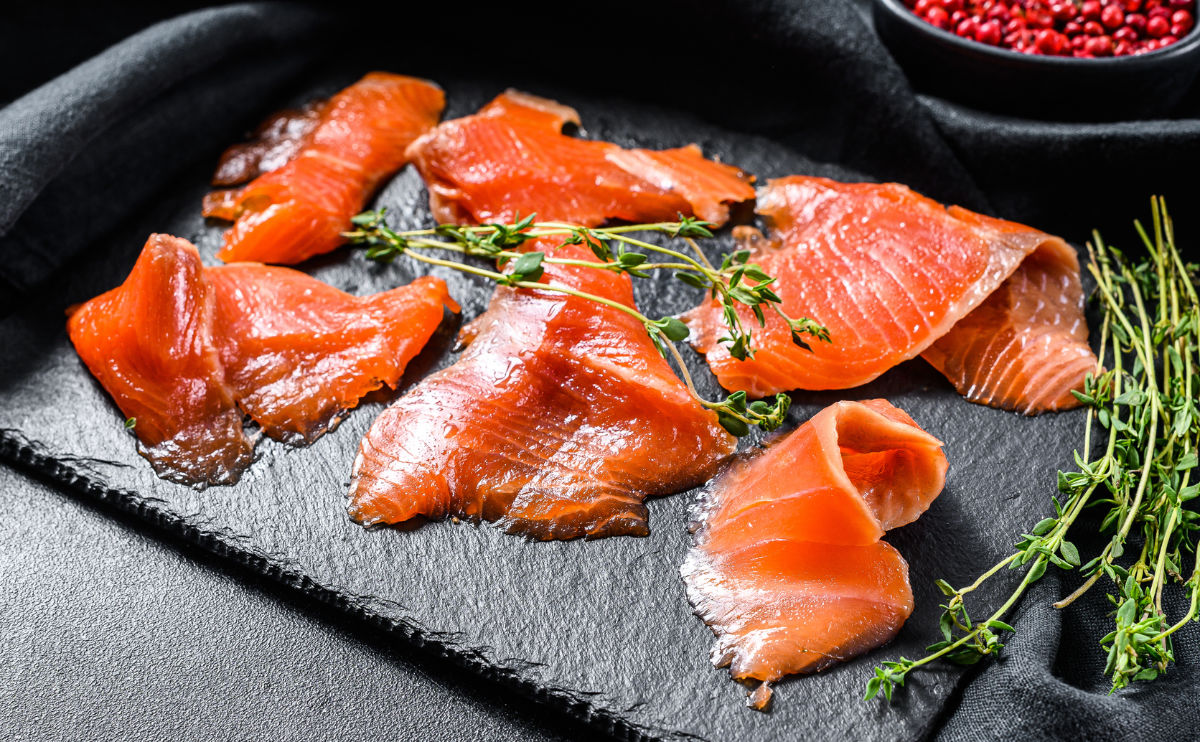 Several slices of smoked trout arranged on a dark slate board, garnished with fresh thyme sprigs. A bowl of red berries sits in the background on a black surface.