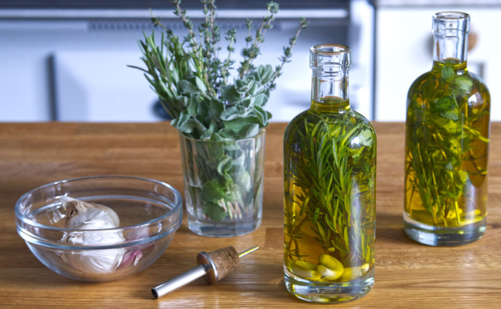 Two glass bottles of herb-infused olive oil with sprigs of rosemary inside sit on a wooden counter. Nearby are a glass bowl with garlic bulbs, a pour spout, and a glass with fresh herbs.