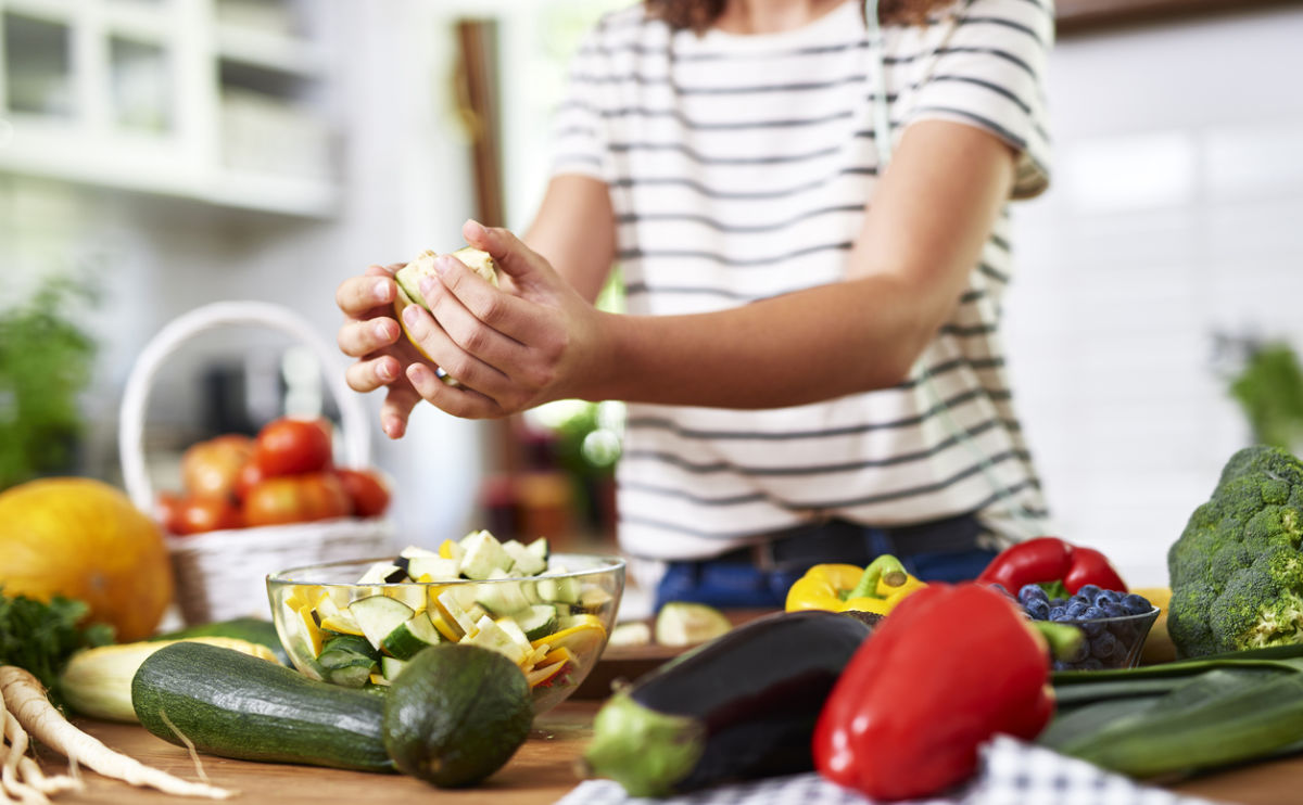 A person in a striped shirt chops vegetables in a bright kitchen, surrounded by fresh produce like zucchini, peppers, broccoli, and tomatoes on a wooden counter.