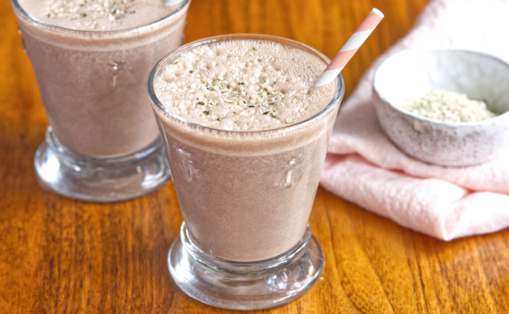 Two glasses of chocolate smoothies with frothy tops sit on a wooden table. One glass has a striped straw. In the background, a small bowl with white seeds rests on a pink cloth.