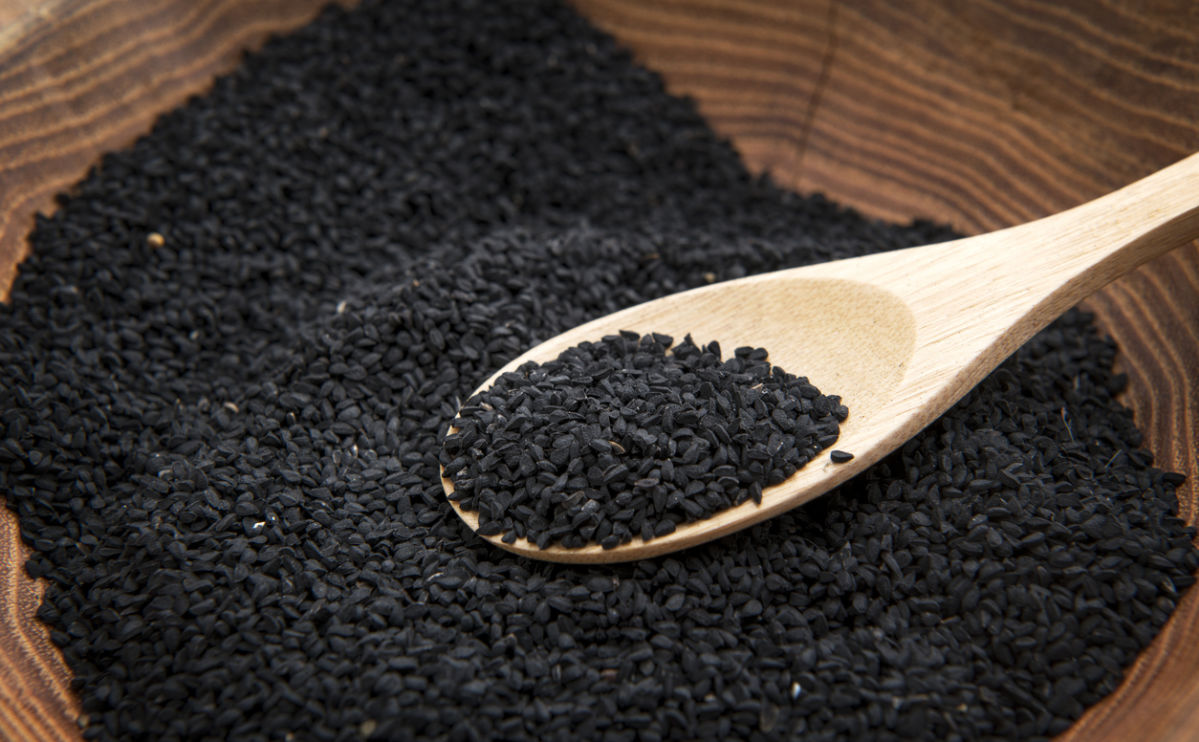 A wooden spoon holding black nigella seeds rests on a wooden bowl filled with more seeds.