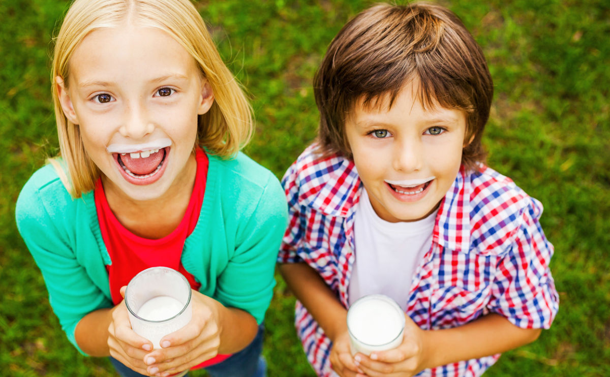 Two smiling children with milk mustaches hold glasses of milk and look up at the camera. They are standing on green grass, and both appear happy and cheerful.