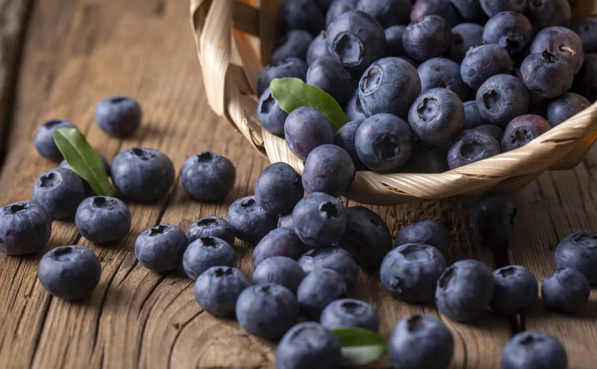 A basket tipped over on a wooden surface with fresh blueberries spilling out, some with small green leaves, creating a natural and rustic scene.