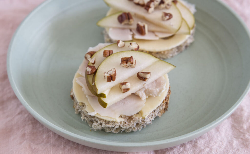 Two round open-faced sandwiches on a light green plate, topped with slices of emmental cheese, chicken, pear, and chopped pecans, served on a light pink tablecloth.