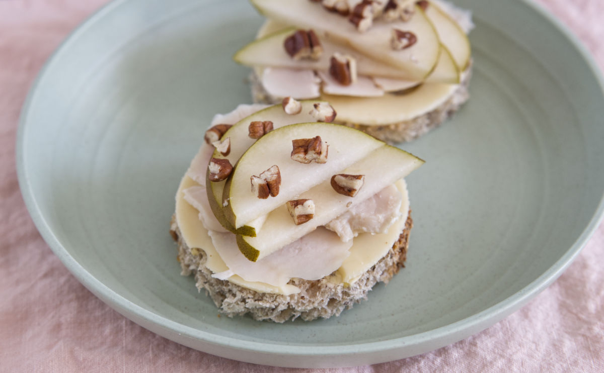 Two round open-faced sandwiches on a light green plate, topped with slices of emmental cheese, chicken, pear, and chopped pecans, served on a light pink tablecloth.