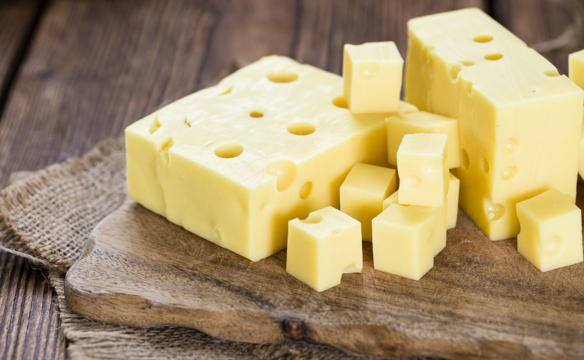 Several blocks and cubes of Emmental cheese with characteristic holes are arranged on a rustic wooden cutting board, placed on a textured burlap cloth over a wooden table.