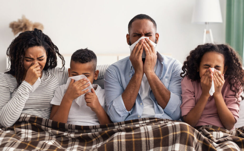 A family of four, sitting on a couch under a plaid blanket, all holding tissues to their noses as if sneezing or suffering from a cold.