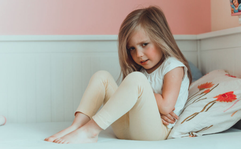 A young girl with long hair sits on a bed, holding her stomach and looking uncomfortable, suggesting she may have a stomach ache. She is wearing a white shirt and light-coloured trousers, with a floral cushion behind her.