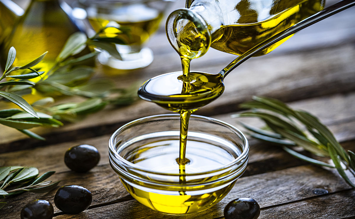 Pouring extra virgin olive oil from a spoon to a glass container. Olive branches comes from the left and right. Some olive oil bottles are out of focus at background. Black olives are on the table and complete the composition. The composition is on a rustic wooden kitchen table. Predominant colors are gold, green and brown. 