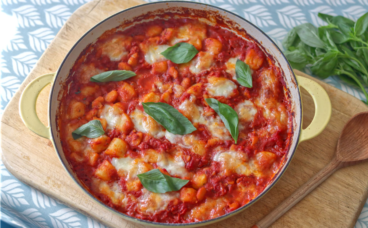 A round baking dish filled with baked gnocchi in tomato sauce, topped with melted cheese and fresh basil leaves, sits on a wooden board next to a wooden spoon and a bunch of basil.