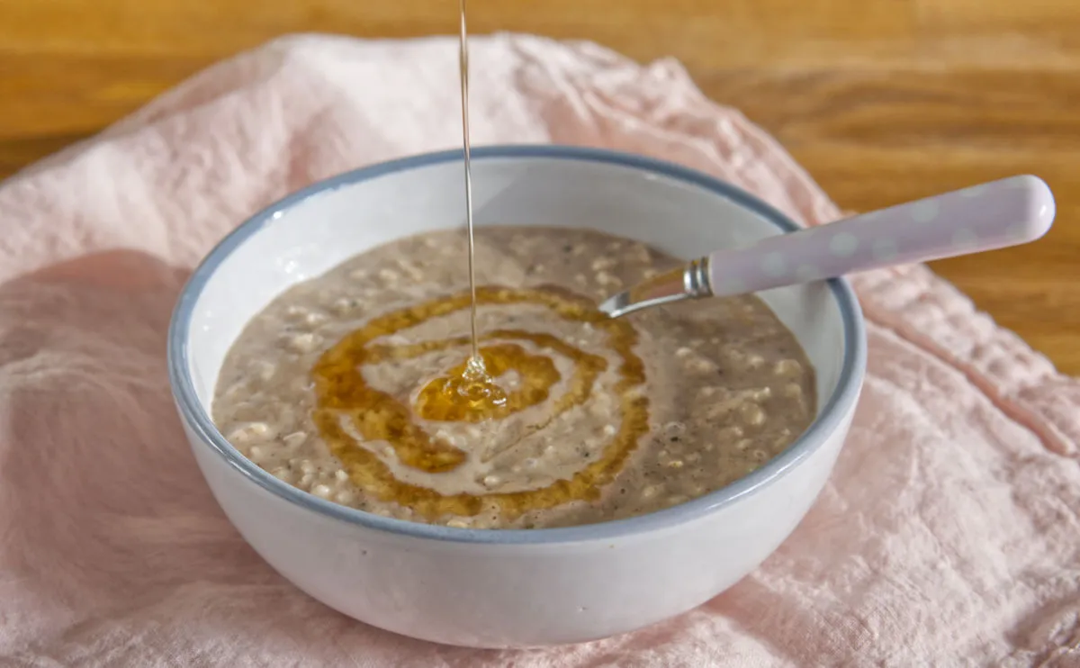 A bowl of porridge with a spoon rests on a pink cloth, while golden honey is being drizzled in a spiral pattern on top.