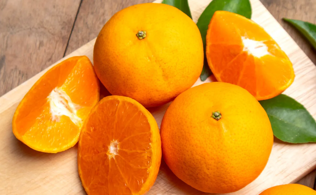 Two whole oranges and three orange halves are displayed on a wooden surface with a few green leaves in the background. The fruit appears fresh and juicy, with vibrant orange colour and smooth rind.