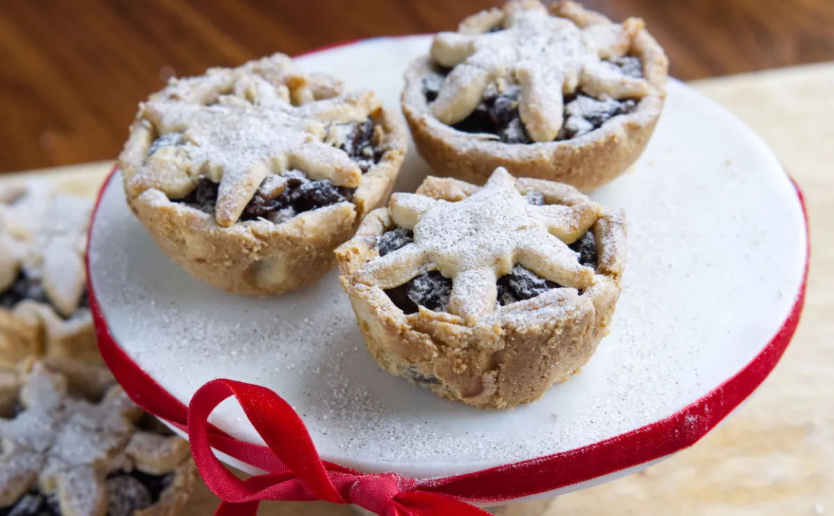 Three individual mince pies with star-shaped pastry tops and a dusting of icing sugar are displayed on a white cake stand decorated with a red ribbon.