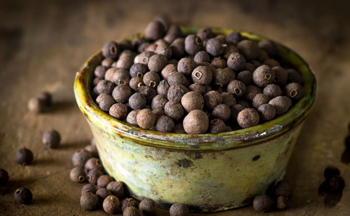 A rustic, green ceramic bowl filled with whole dried allspice berries sits on a wooden surface, with a few loose berries scattered nearby.