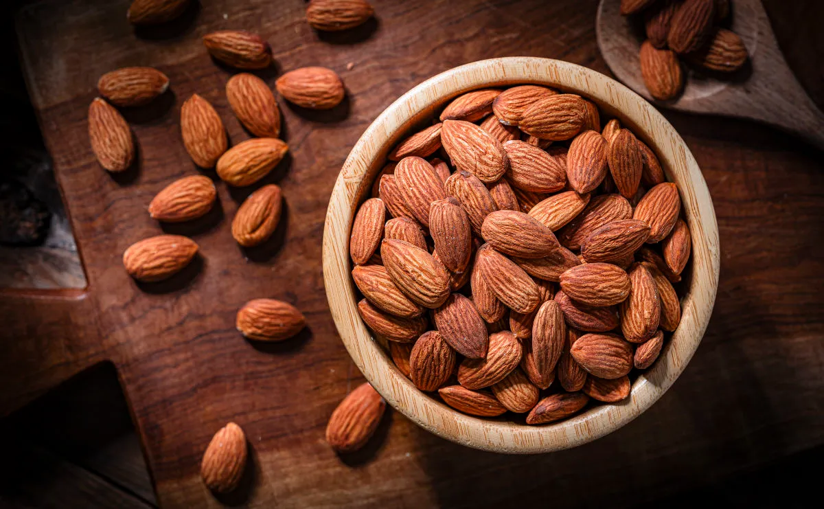 A wooden bowl filled with whole, raw almonds sits on a wooden surface, with more almonds scattered around it.