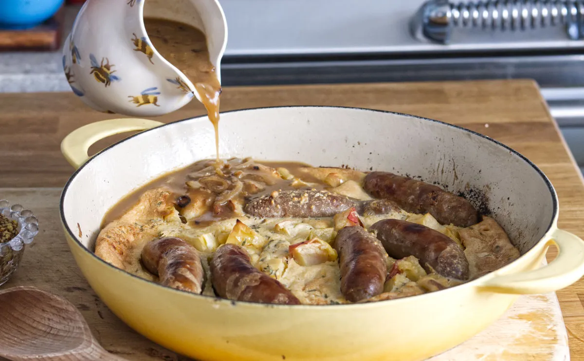 Pouring gravy over sausages and mash in yellow pan