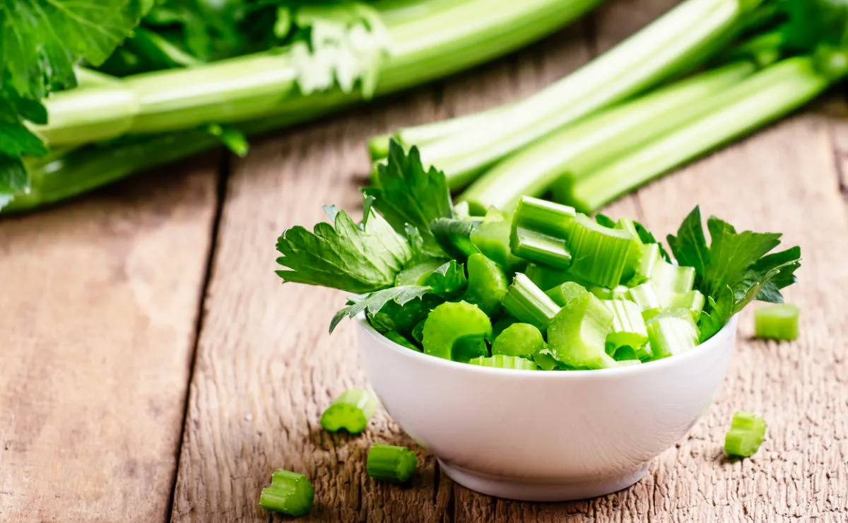 A white bowl filled with chopped celery sits on a wooden surface, with fresh celery stalks and leaves in the background.