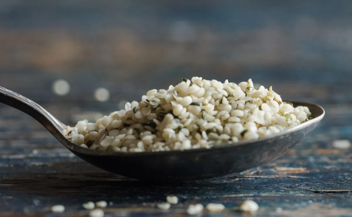 A close-up of a spoon filled with hemp seeds, resting on a rustic wooden surface. The seeds are small, pale, and slightly greenish in colour.