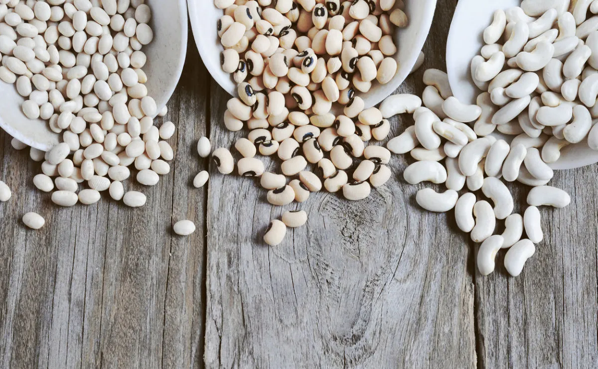 Three piles of beans—haricot beans, black-eyed peas, and cannellini beans—are arranged on wooden spoons atop a rustic wooden surface, with some beans scattered around.