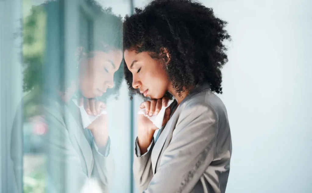 Woman looking sad and anxious, leaning her head against a window with her face reflected back at her.