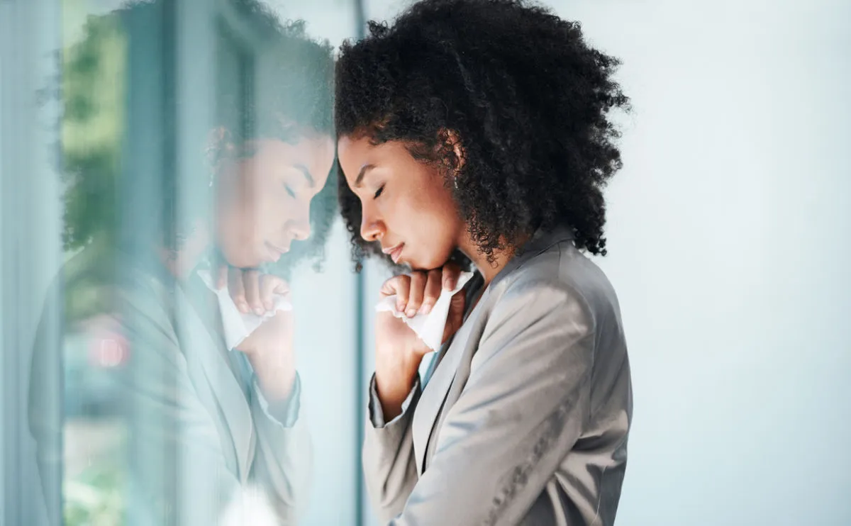 Woman looking sad and anxious, leaning her head against a window with her face reflected back at her.