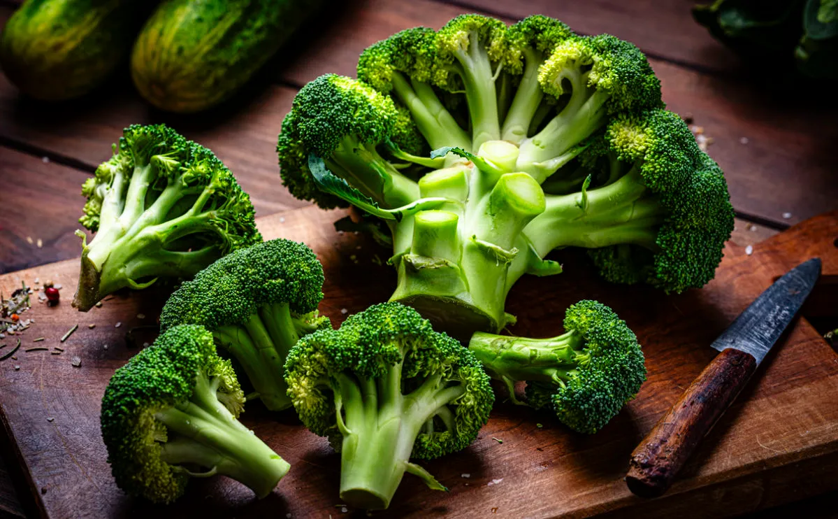 head of broccoli on a wooden chopping board, with a few florets cut off across the chopping board.