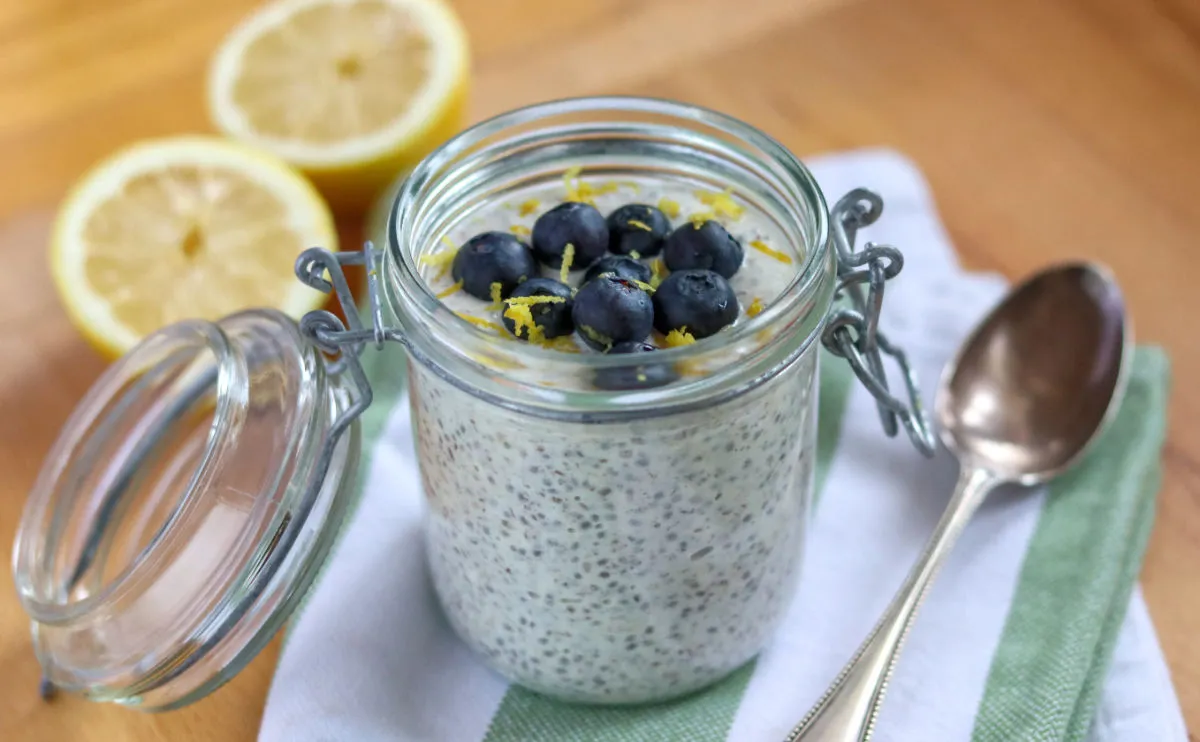 Glass jar filled with yoghurt and chia seeds, blueberries and lemon zest are sprinkled on top and there are lemons in the background.