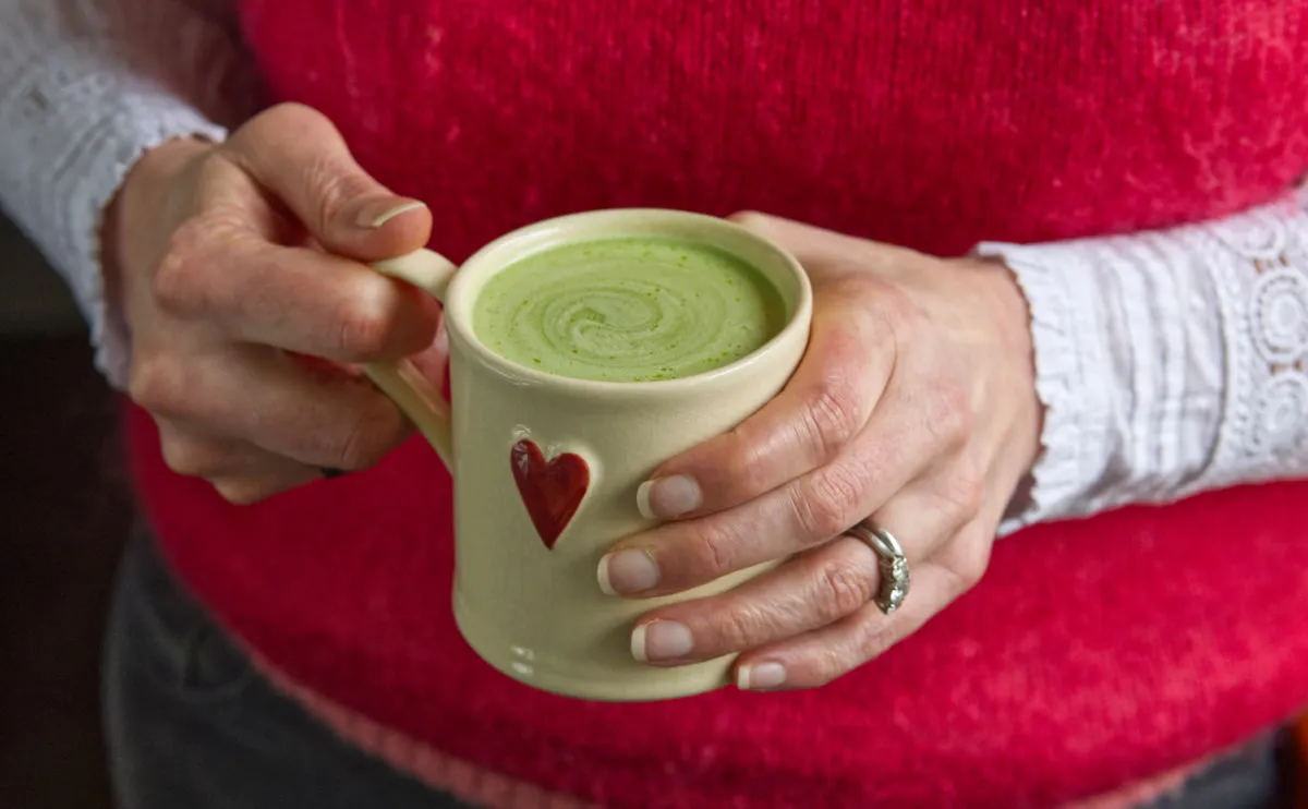 A lady in a red top is holding a cream cup full of Matcha tea, swirled with milk.