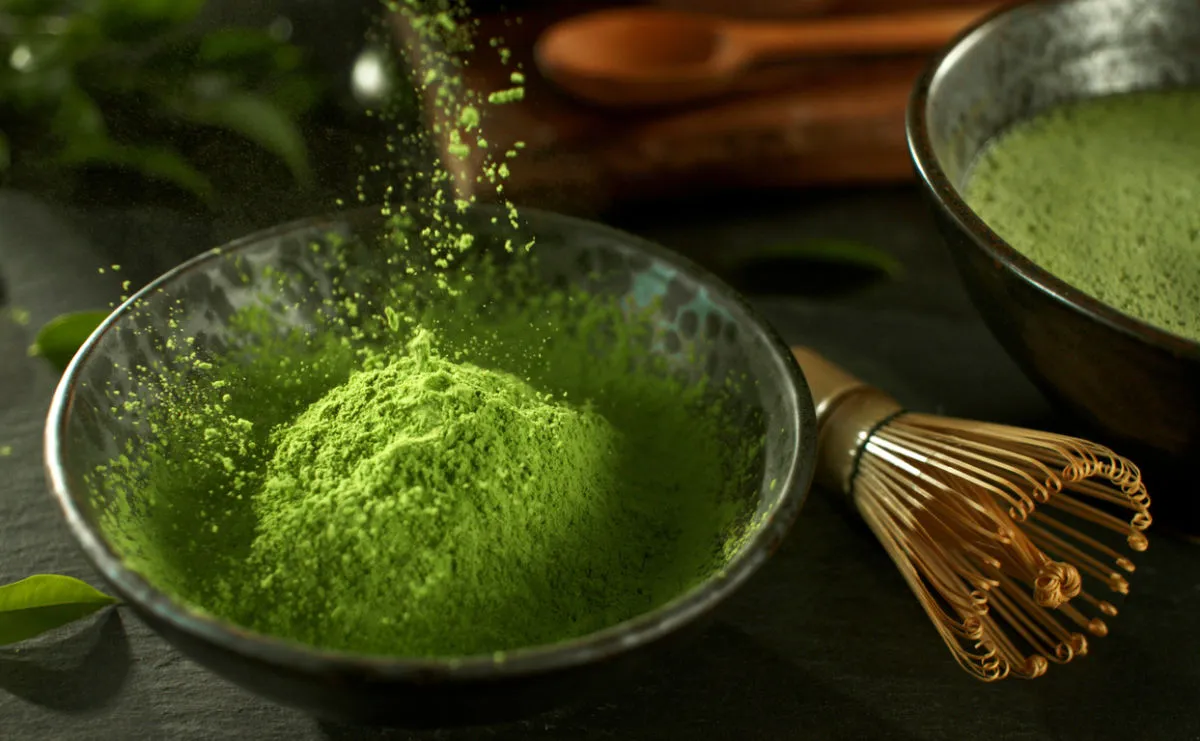 Matcha Green tea powder in a bowl, next to a bamboo whisk and a bowl of matcha tea. There are tea leaves scattered around the bowl.