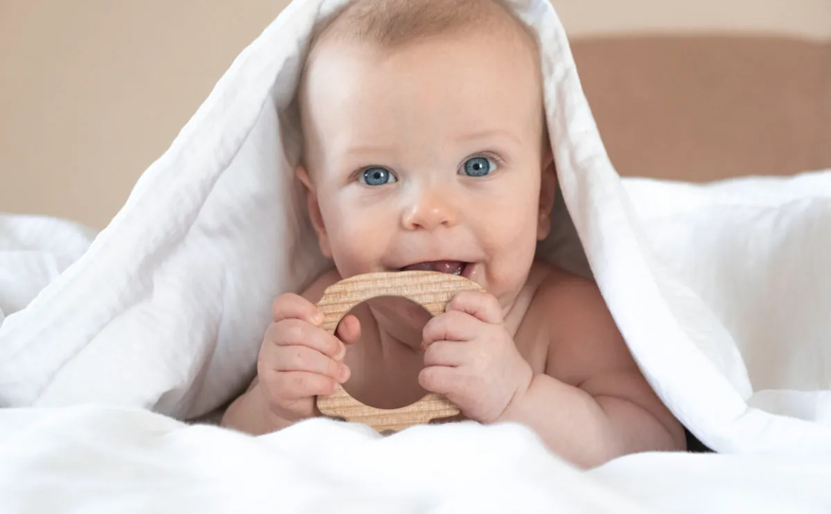Baby with a white towel over its head, peaking out with a wooden teether held to their mouth.