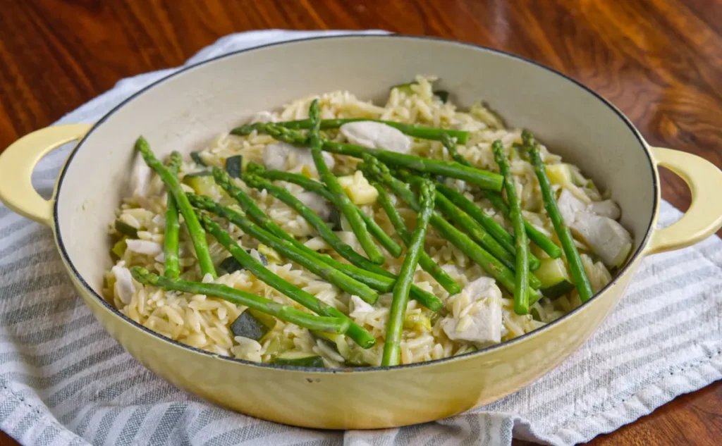chicken, orzo and asparagus in a yellow casserole dish on top of a blue and white striped tea cloth