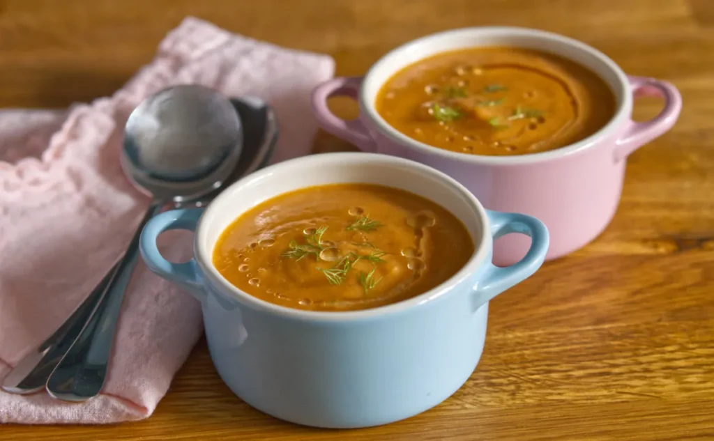 Tomato soup in pink and blue bowls next to a pink napkin with a silver spoon.