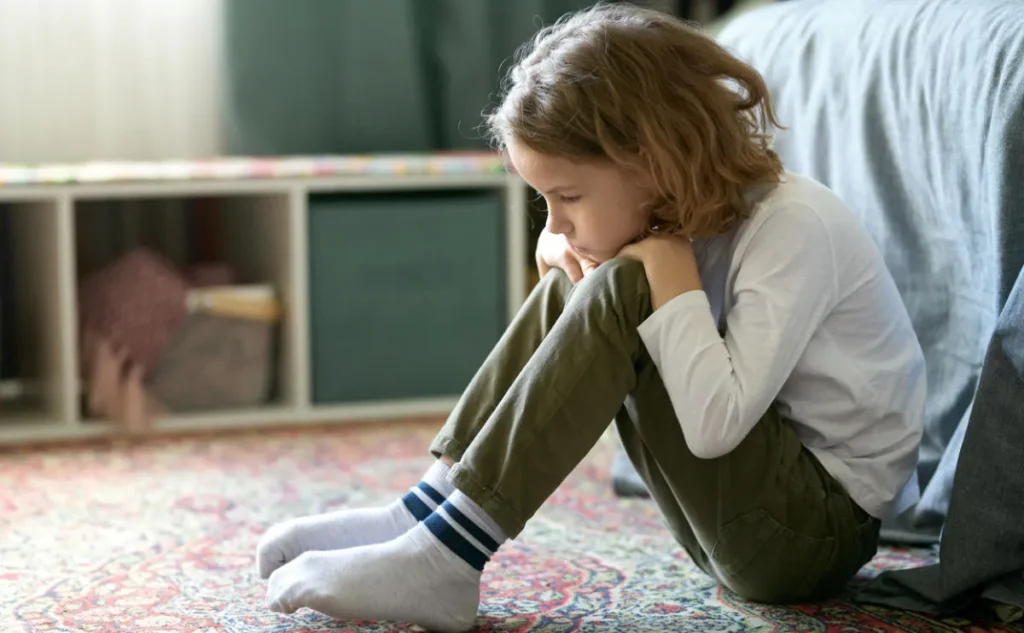 Young girl looking sad and anxious, holding her knees up to her chest by her bed.