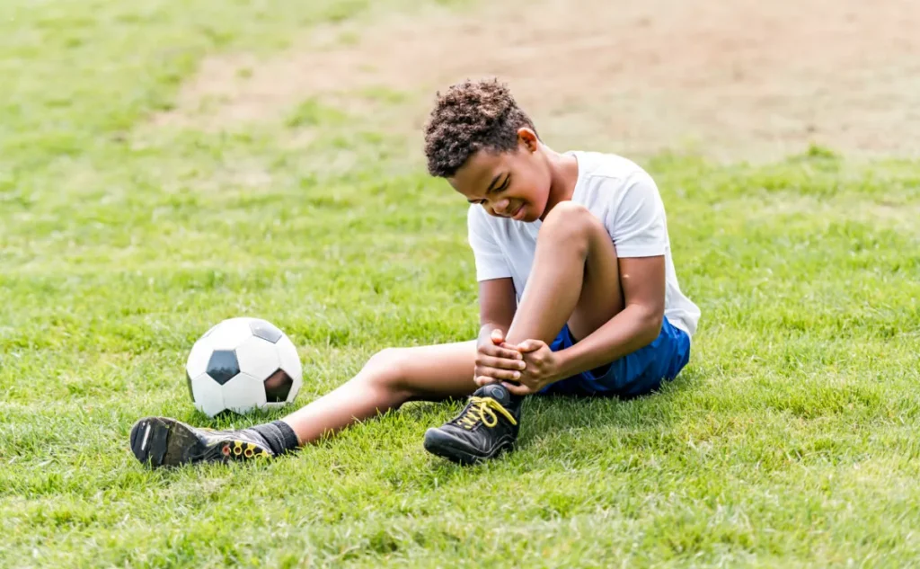 Boy in football kit, sitting next to a football, clasping his ankle in pain.