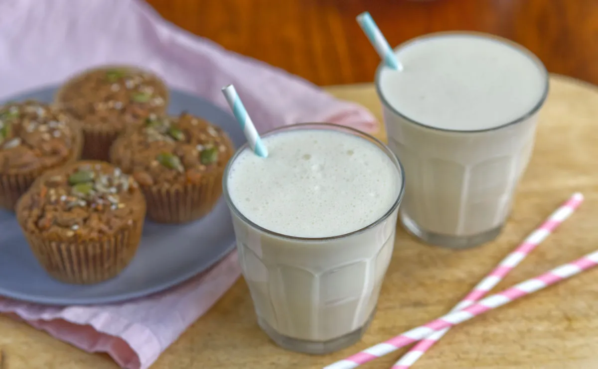 Two glasses of vanilla milkshake with blue and white striped straws, next to a plate of muffins.