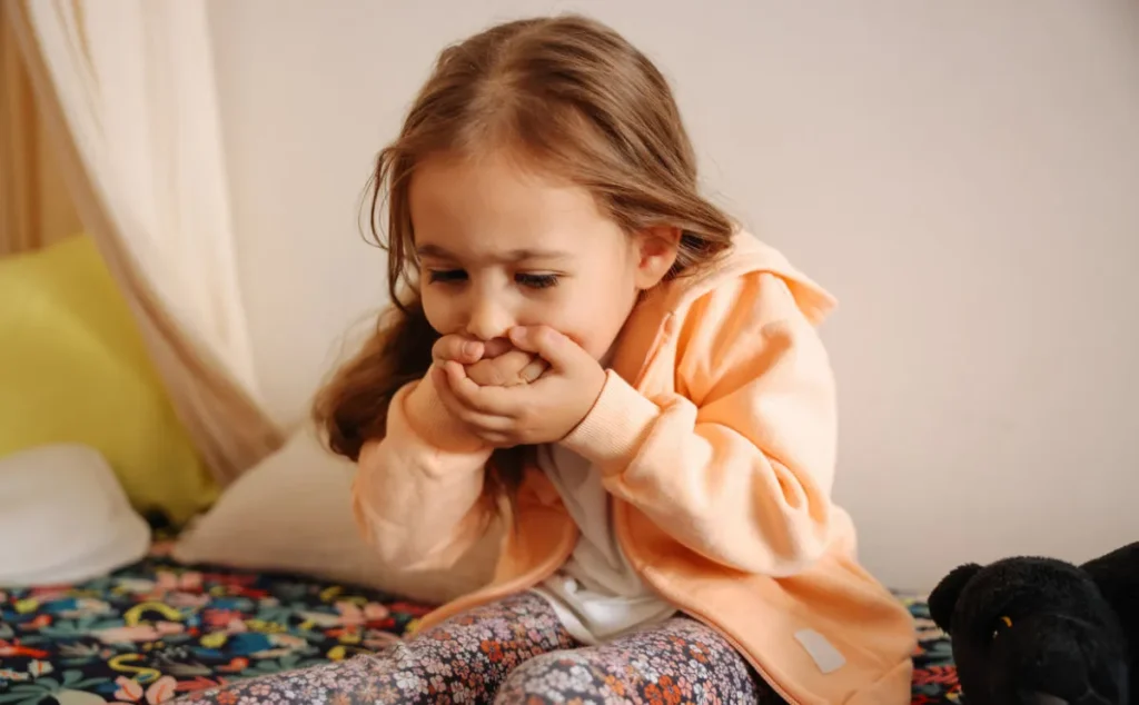 Little girl in apricot coloured dressing gown, holding her hands over her mouth as if she is about to be sick