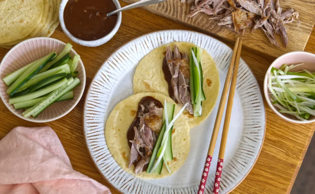 A white plate with two pancake style wraps with duck and spring onions, bowls of cucumber, spring onion and hoisin sauce surround the plate.