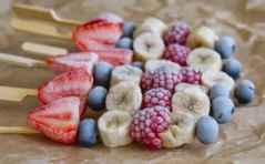 Photograph of frozen fruit, strawberry, blueberry, banana and raspberry all glistening from the freezer, threaded on bamboo skewers.