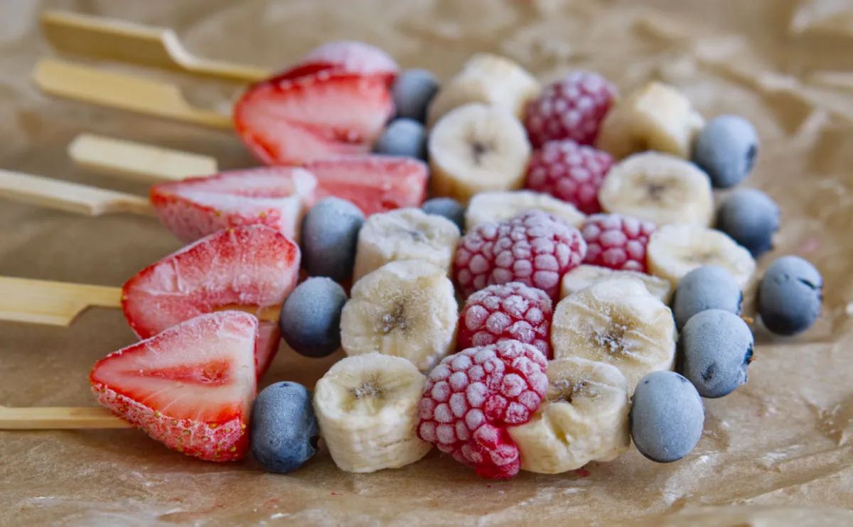 Photograph of frozen fruit, strawberry, blueberry, banana and raspberry all glistening from the freezer, threaded on bamboo skewers.