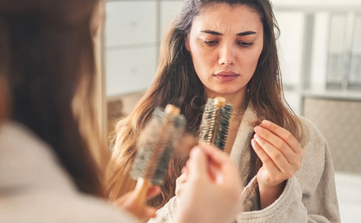 Woman with long brown hair, looking at her hairbrush and noticing a lot of her hair has come out. She is reflected in a mirror.