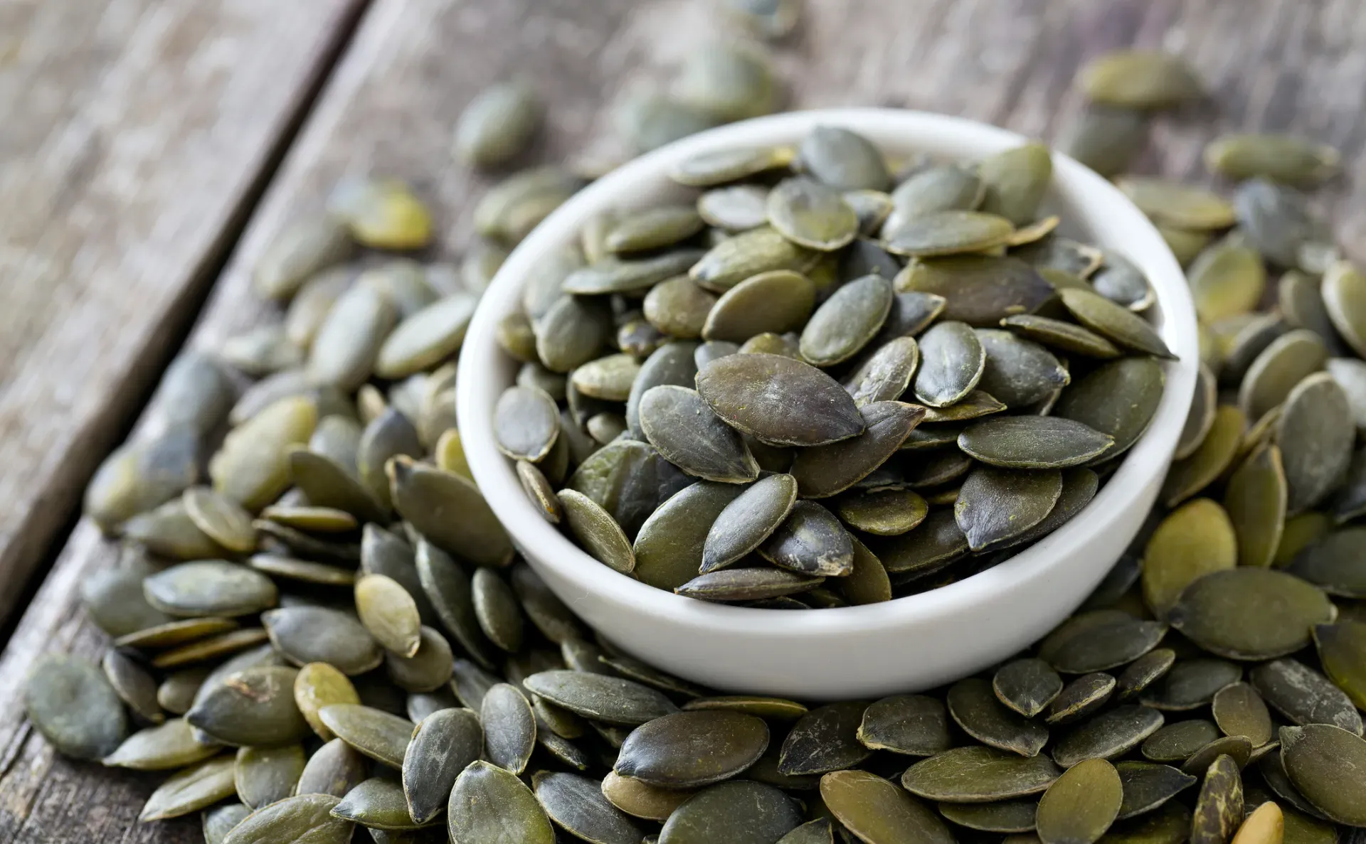 Small white bowl full of pumpkin seeds, which are overflowing onto wooden boards.
